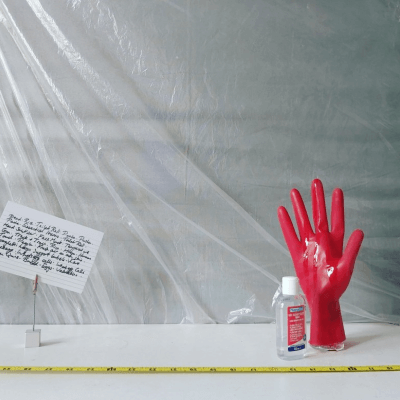 A photograph of a red rubber glove, a small bottle of liquid, a hand written note and a tape measure on a table. The wall behind is covered in clear plastic.